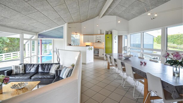 En rummelig opholdsstue med et moderne køkken, et spiseområde med et langt bord og en siddeplads med sofa og stole.