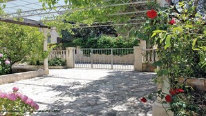 Une cour dallée de pierres, fermée par un portail en métal, et entourée d’une végétation luxuriante et de plantes en fleurs.