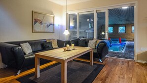 A living room with a black leather sofa, a wooden coffee table, and a painting on the wall. There is a glass door leading to a pool area. - 5 bedroom stunning home in Fredericia (Fredericia)