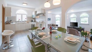 Een moderne keuken met een eethoek, met een glazen tafel, groene stoelen en een televisie op de achtergrond.