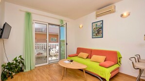 A cozy living room with a wooden floor, a green and orange sofa, a small round table, and a sliding glass door leading to an outdoor balcony. - 2 bedroom beach front apartment in Makarska (Makarska)
