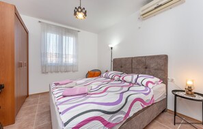 Ein Schlafzimmer mit Bett, Nachttischlampe, Fenster mit leichtem Vorhang, Kleiderschrank und Klimaanlage.