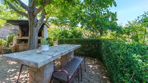 Una mesa de piedra al aire libre con bancos, un árbol y un seto al fondo.