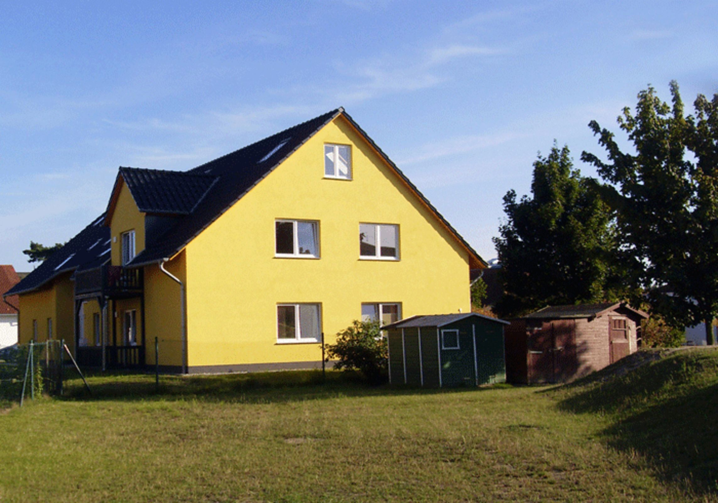 A yellow two-story house with a dark roof, a small shed, and a grassy yard.