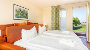 Hotellihuone, jossa on suuri sänky, oranssit ja valkoiset tyynyt, taulu seinällä ja näkymä vehreään maisemaan ikkunasta.
