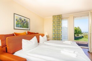 A hotel room with a large bed, orange and white pillows, a painting on the wall, and a view of greenery through the window. - Ferienapartment-ruegen - 2-room apartment (Sagard)