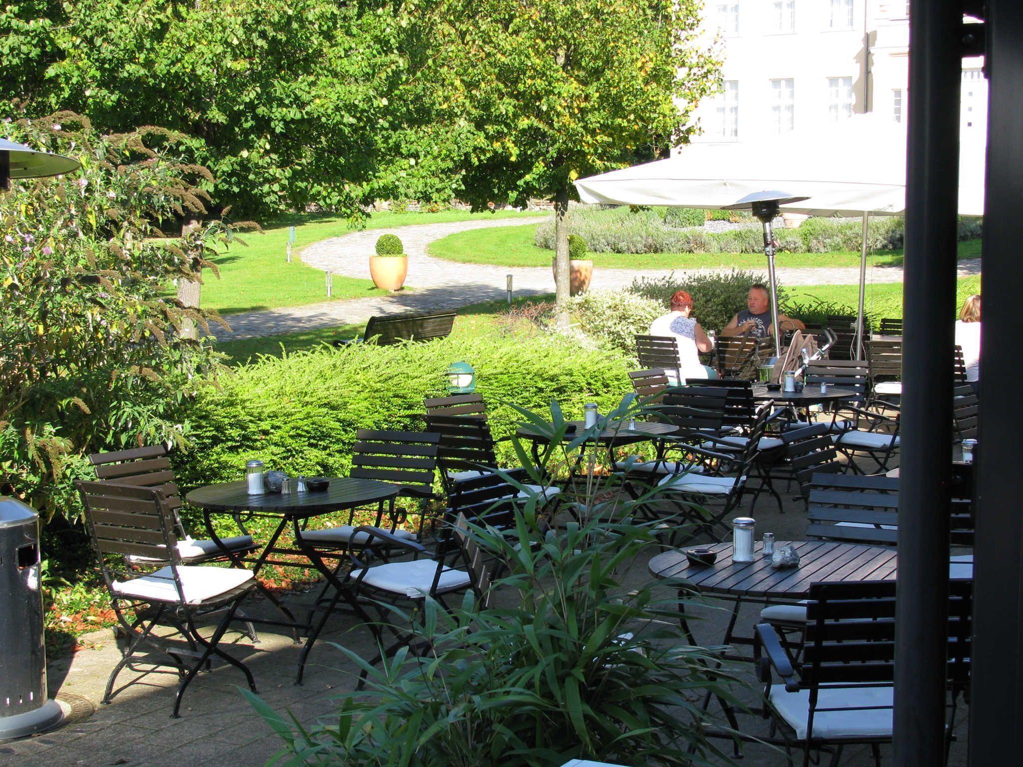 An outdoor patio with tables and chairs, surrounded by greenery and a pathway leading to a building.