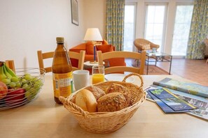 A breakfast table with a basket of bread, a bottle of juice, a bowl of fruit, and a magazine.