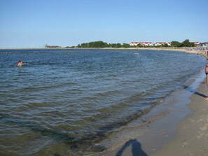 A beach with clear water, a sandy shore, and buildings in the background.