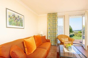 A living room with a sofa, a wicker chair, a glass coffee table, and a view of the outdoors. - Ferienapartment-ruegen - 2-room apartment (Sagard)