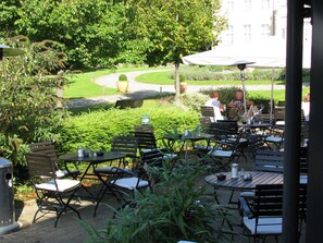 Eine Terrasse im Freien mit Tischen und Stühlen, umgeben von Grünpflanzen und einem Weg zu einem Gebäude.
