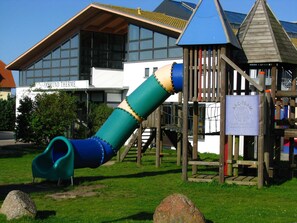 A playground with a slide and climbing structures, set against a building with large windows and a sign reading "HASMUND THERMIC". - Ferienapartment-ruegen - 2-room apartment (Sagard)