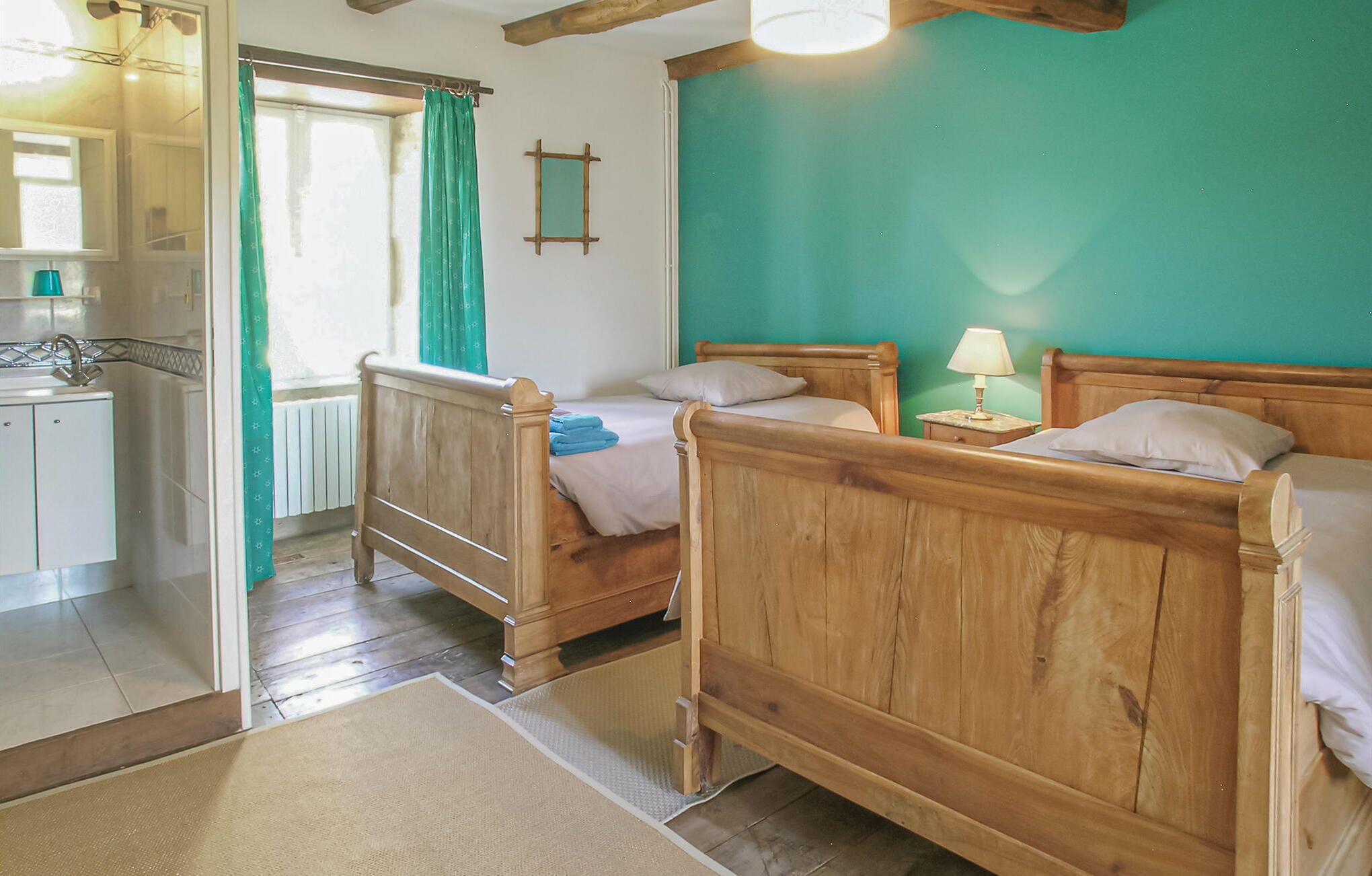 Une chambre à coucher avec deux lits en bois, une petite table de chevet avec une lampe, une fenêtre avec des rideaux et une salle de bain avec un lavabo et un miroir.