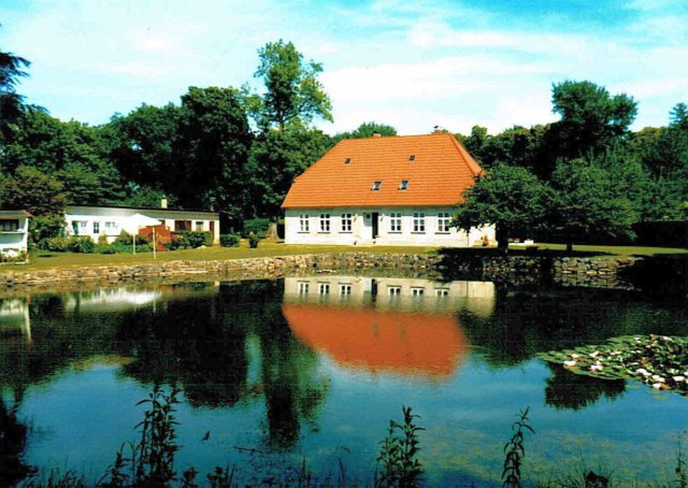 Ein Haus mit rotem Dach, einem Teich im Vordergrund und Bäumen im Hintergrund.