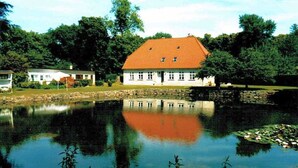 Ein Haus mit rotem Dach, einem Teich im Vordergrund und Bäumen im Hintergrund.