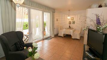 Ein Wohnzimmer mit einer Couch, einem Esstisch mit Stühlen, einem Fernseher und einem Balkon mit Vorhängen.