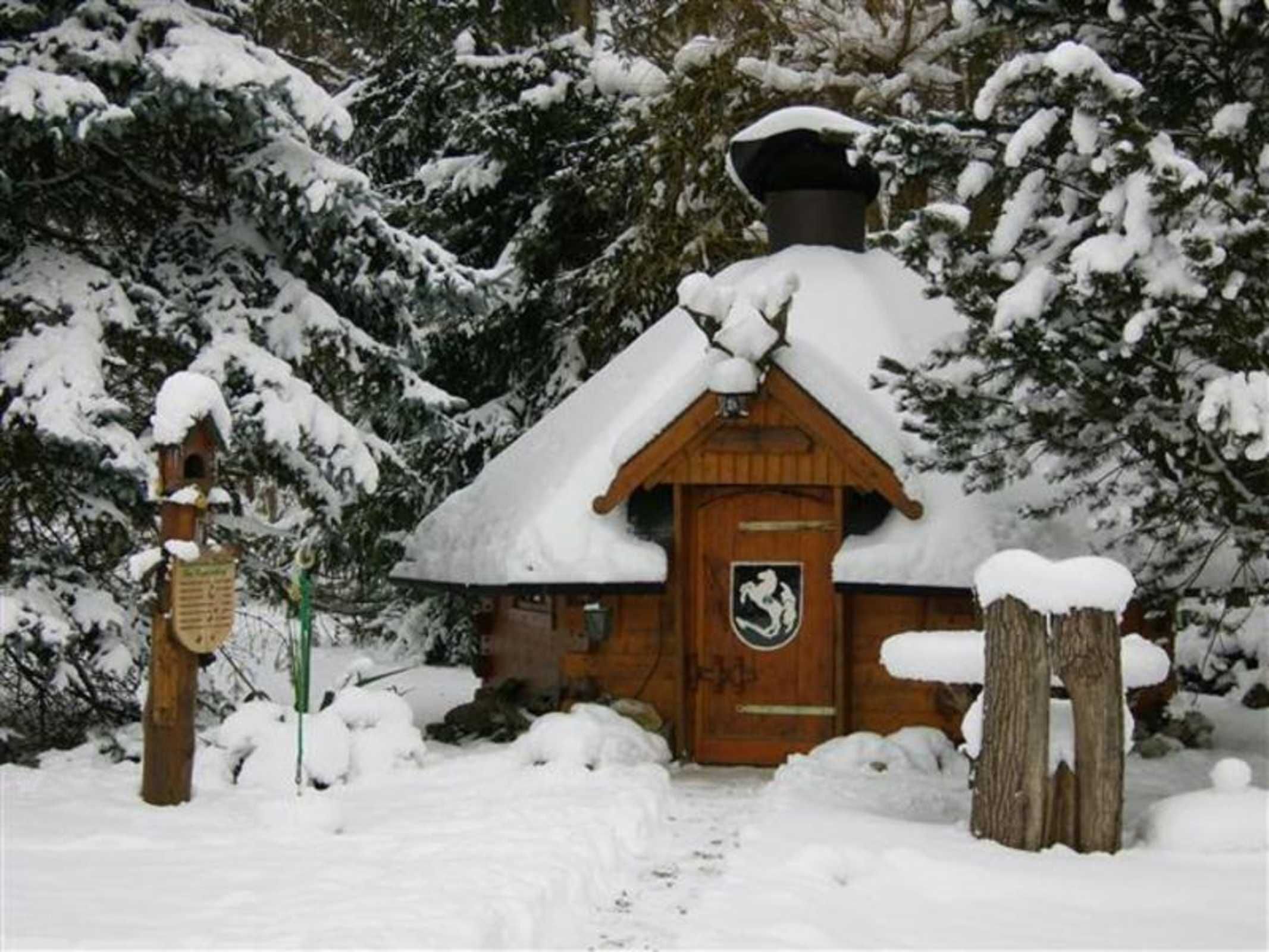 Eine Holzhütte mit schneebedecktem Dach, eine Holztür mit Wappen und ein Holzpfosten mit einem Schild.