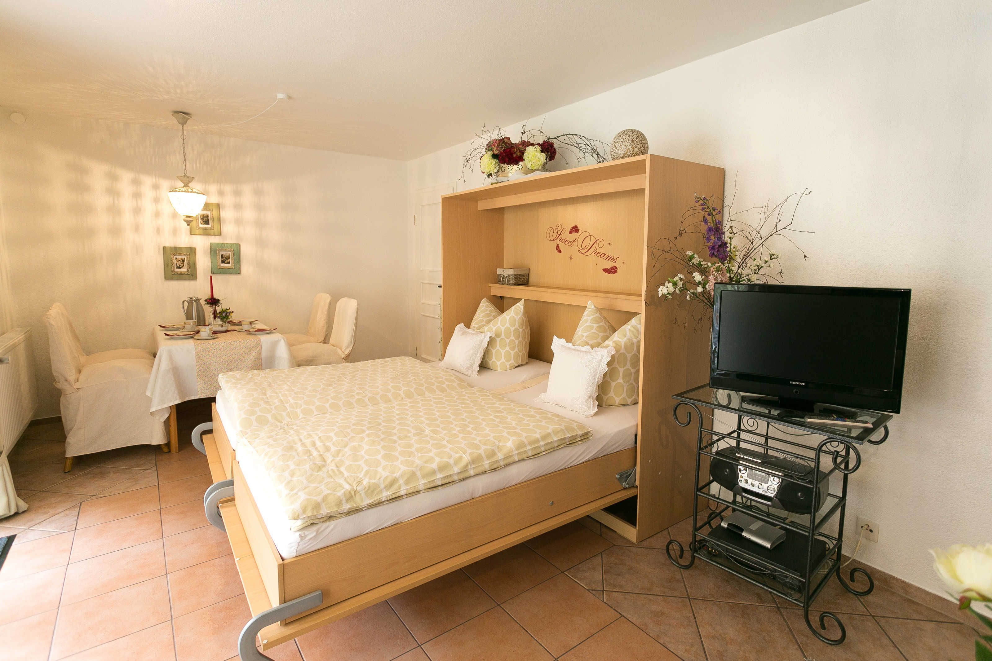 A bedroom with a bed, a TV stand with electronics, a bedside table, and a chair.