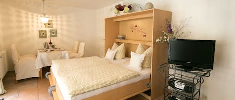 A bedroom with a bed, a TV stand with electronics, a bedside table, and a chair.