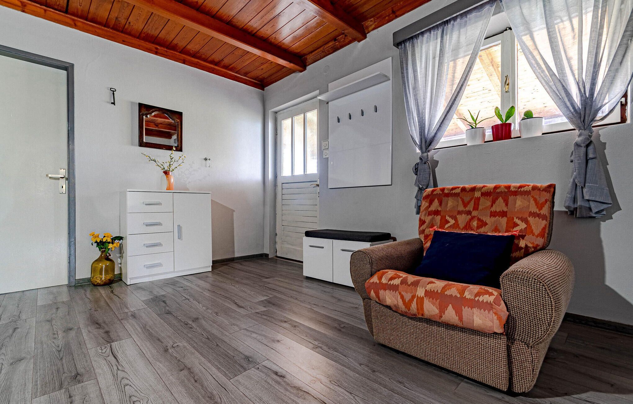 A room with a wooden ceiling, a white cabinet with drawers, a wicker chair with a patterned cushion, and a window with sheer curtains.