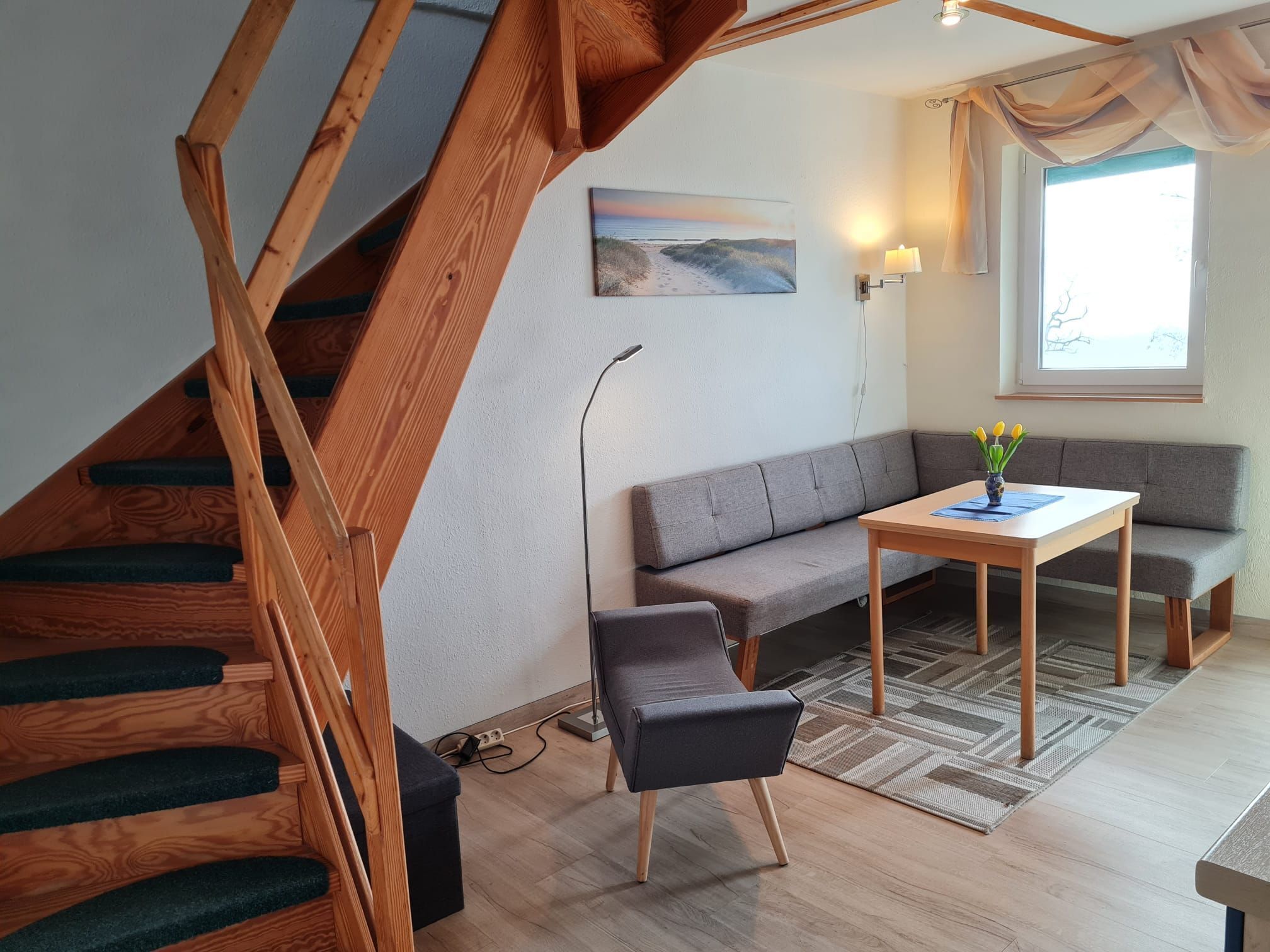 A modern living room with a wooden staircase, grey sofa, and a small table with a vase.
