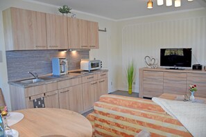 A modern kitchen with wooden cabinets, a built-in microwave, and a toaster. There is a dining area with a striped sofa and a small table. A TV is mounted on the wall above a wooden dresser.