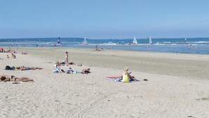 Una playa arenosa con gente tomando el sol y nadando, y veleros a lo lejos.