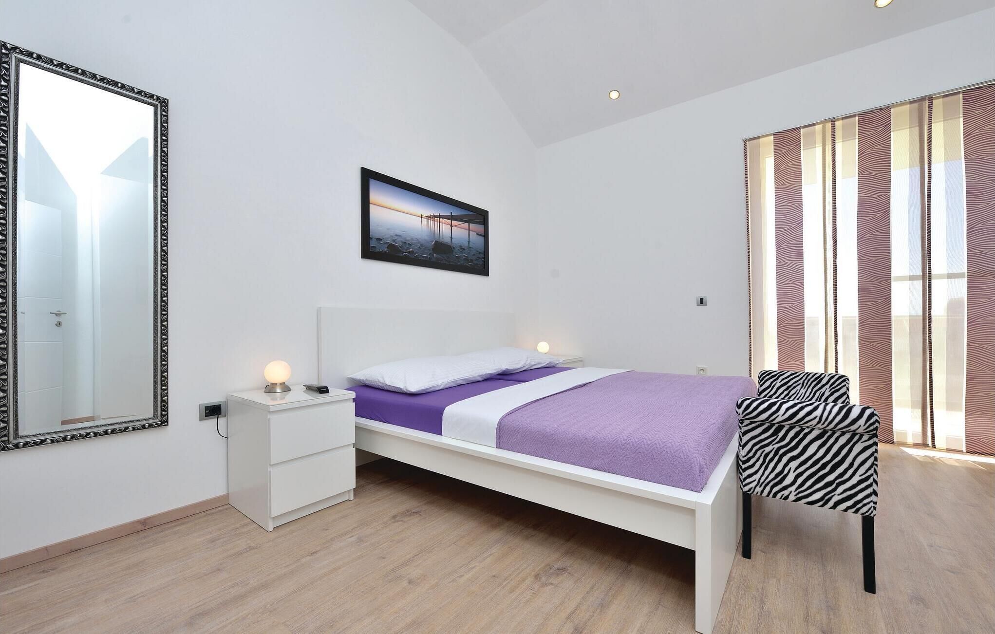 A bedroom with a bed, a nightstand, a chair, a large mirror, and a picture on the wall.
