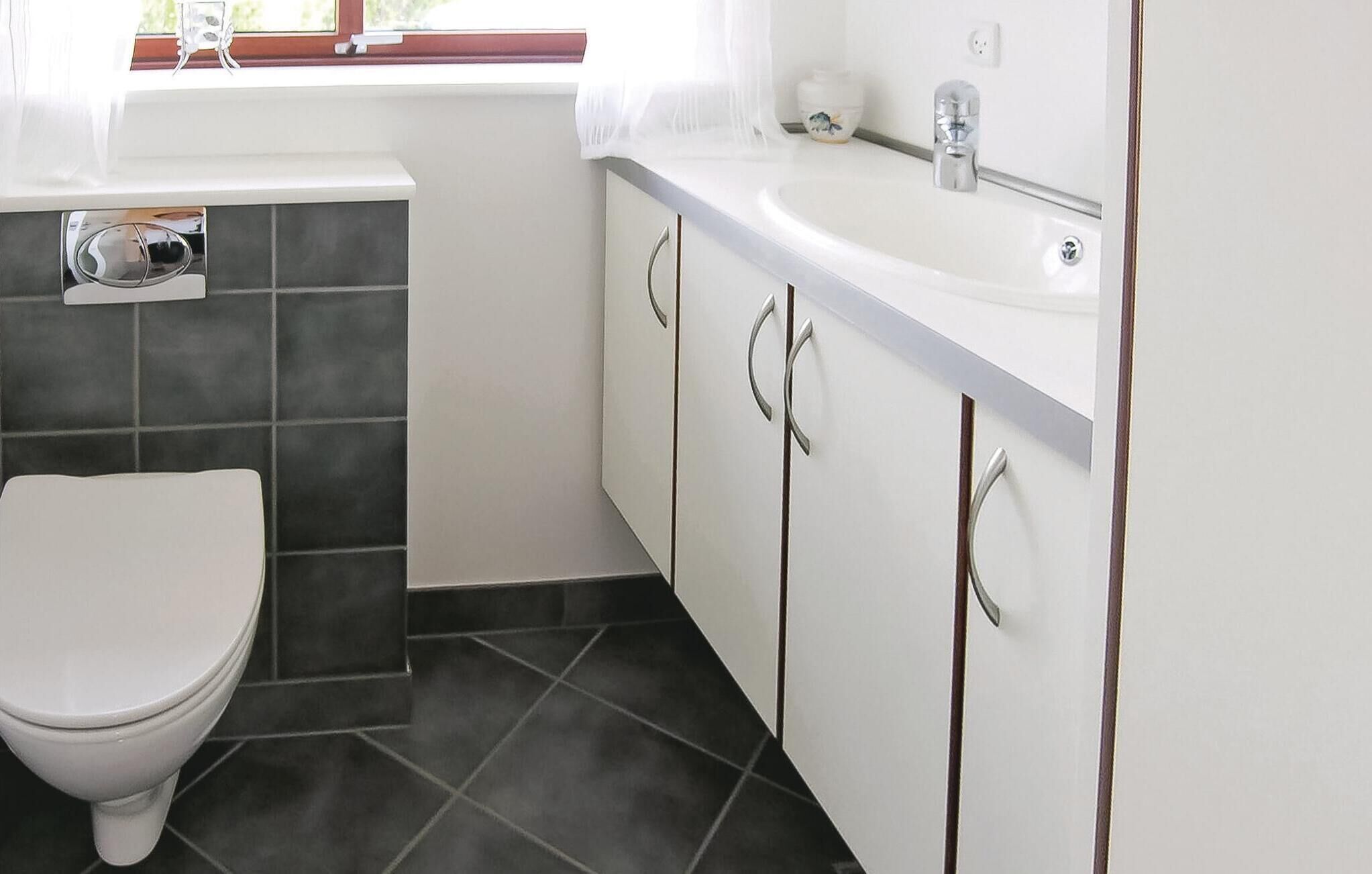 Une salle de bain avec des toilettes, un carrelage foncé, des meubles blancs et une fenêtre ornée de rideaux légers.