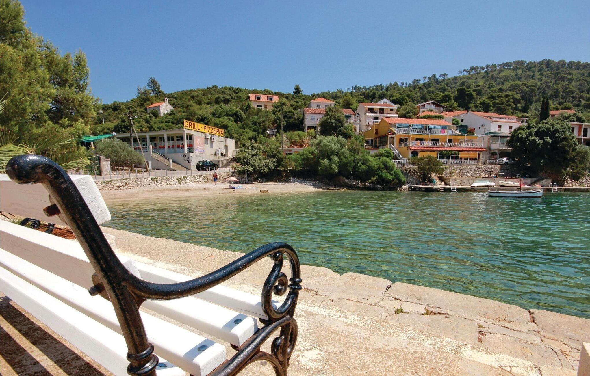 A coastal view with a bench overlooking a calm sea, a building labeled 'GRILL - PIZZERIA', and a row of houses along the hillside.