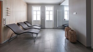 Une salle d’attente comprenant trois fauteuils noirs, un mur recouvert de panneaux en bois, un radiateur et une fenêtre avec des stores.