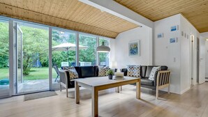 A spacious living room with a wooden ceiling, large sliding glass doors leading to a garden, a central table, and a sofa with patterned cushions. - Lovely home in Oksbøl with sauna (Oksbøl)
