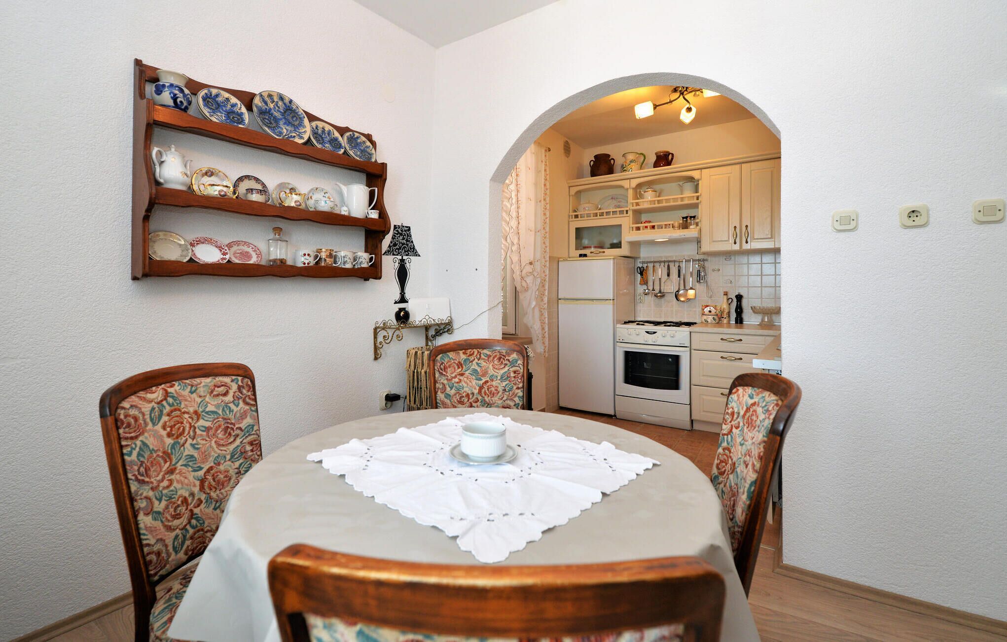 A dining area with a round table, floral chairs, and a wall-mounted shelf with dishes.