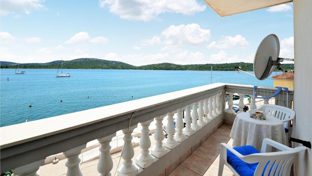 Un balcón con vista al mar tranquilo, una barandilla blanca, una mesa con una silla y una antena parabólica.