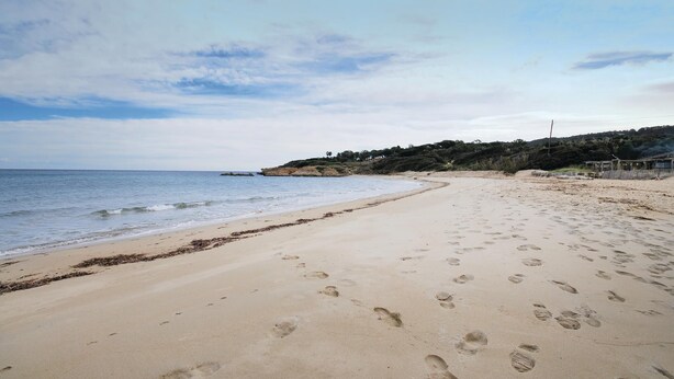 Une vaste plage de sable où l’on peut voir des empreintes de pas menant à l’eau, une mer calme et un paysage vallonné avec une végétation clairsemée en arrière-plan.