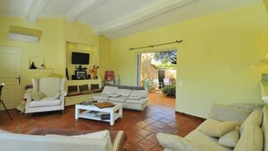 Un salon avec un canapé blanc, un fauteuil blanc et une table basse. Un téléviseur est fixé au mur et une porte donne accès à un espace extérieur.