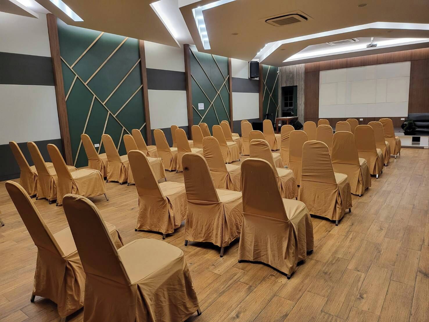 Uma sala de conferências com fileiras de cadeiras cobertas com capas bege, piso de madeira e uma tela grande na frente.