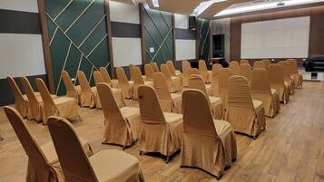 Uma sala de conferências com fileiras de cadeiras cobertas com capas bege, piso de madeira e uma tela grande na frente.