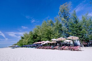 Ein Strand mit Reihen von Liegestühlen und Sonnenschirmen, einem klaren Himmel und dichter Baumlandschaft im Hintergrund.