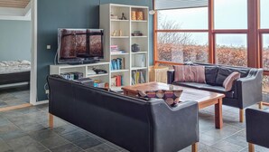 A living room with a black sofa, wooden coffee table, and a TV on a white stand. There is a bookshelf with various items and a window with a view of trees.
