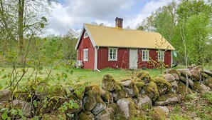 Une maison en bois rouge, avec un toit jaune, entourée de verdure et d’un mur de pierres.