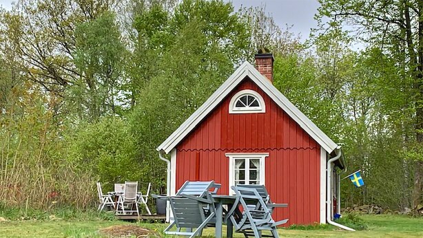 Une maison en bois rouge, avec une fenêtre et une porte blanches, entourée de verdure et d’un drapeau.