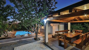 Un espace piscine comprenant des chaises longues, une terrasse en bois et un coin salon couvert avec des tables et des bancs.