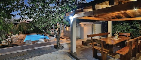 Área de piscina com espreguiçadeiras, deck de madeira e área de estar coberta com mesas e bancos.