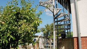 A metal spiral staircase attached to a building, surrounded by greenery and a fence. - Vacation apartment with garden terrace - Holiday apartments with terrace (Patzig)