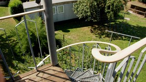 A residential setting with a metal staircase leading to a deck, surrounded by greenery and a small house. - Vacation apartment with garden terrace - Holiday apartments with terrace (Patzig)