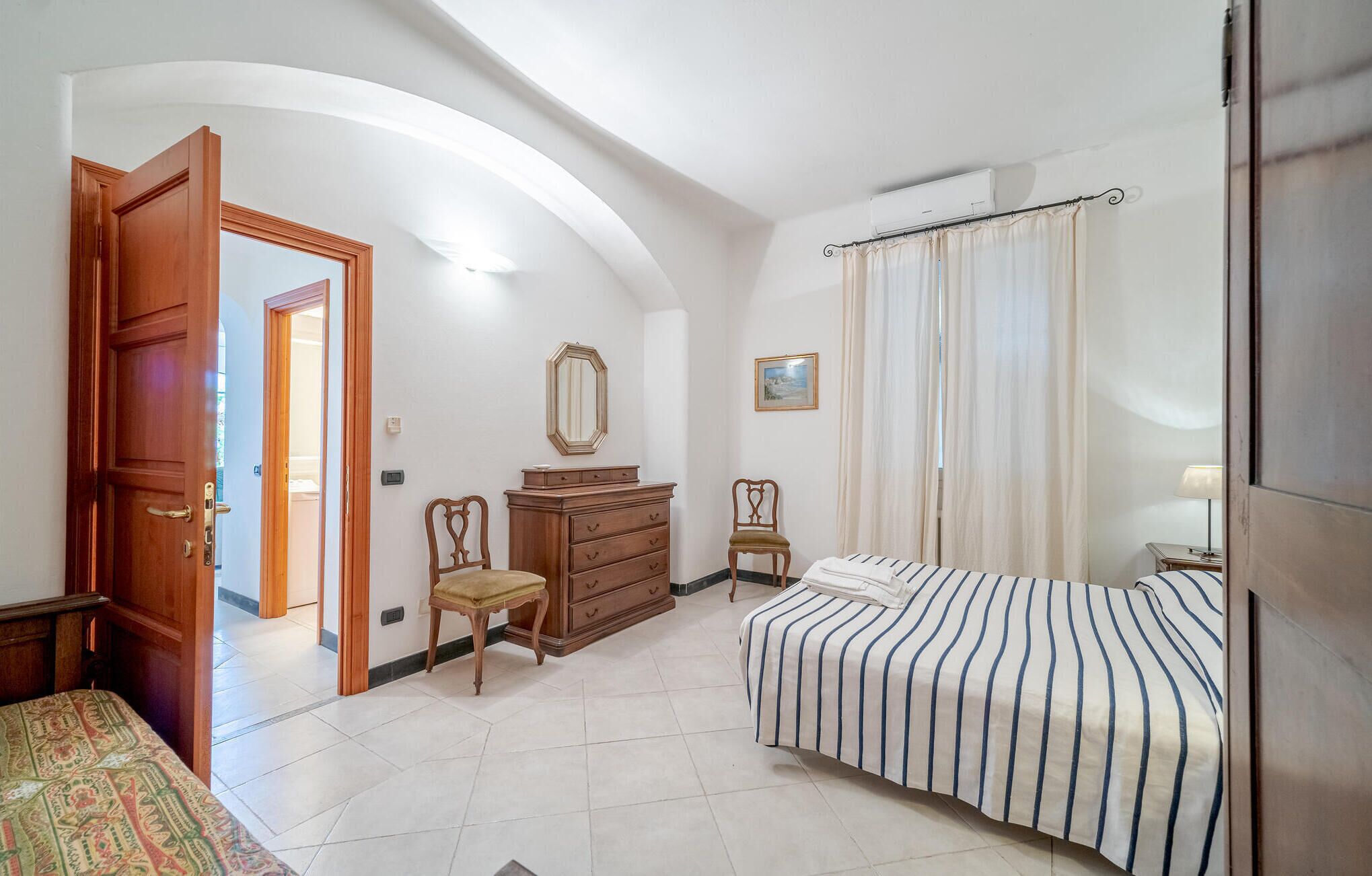 A bedroom with a bed, a dresser, a chair, a mirror, and a door leading to another room.