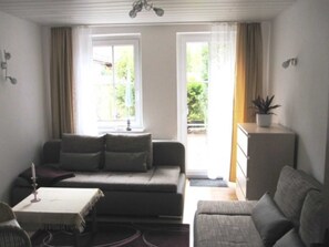 A living room with a sofa, a small table, and a cabinet. There are two windows with curtains, and a plant on the cabinet.