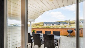 Eine überdachte Terrasse mit Korbmöbeln und einem Grill, mit Blick auf eine Ziegelmauer und ein Gebäude mit Solaranlagen.