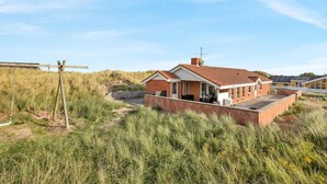 Ein eingeschossiges Haus mit Backsteinfassade und Ziegeldach, umgeben von hohem Gras und Dünen.